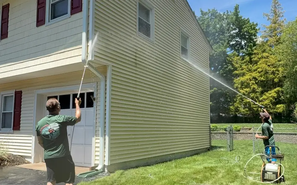 Two workers pressure washing the vinyl siding of a two-story house.